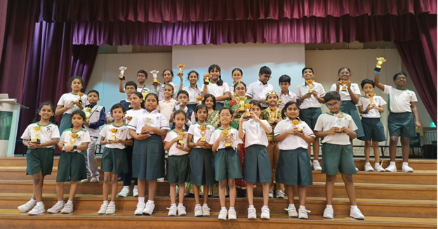 A large group of school children in uniforms pose on a stage holding trophies and medals.